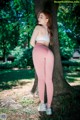 A woman in a white top and pink leggings leaning against a tree.