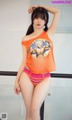 A woman in an orange shirt and pink panties posing for a picture.