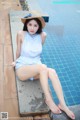 A woman in a blue dress and straw hat sitting by a swimming pool.